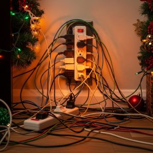 an overloaded wall outlet with too many plugs, tangled cords, and a faint glow of overheating near Christmas decorations.